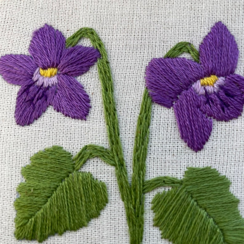 May include: Embroidered purple violets with green stems and leaves on a white linen background.