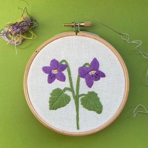 May include: A purple and green embroidered flower design on a white fabric stretched in a wooden embroidery hoop.