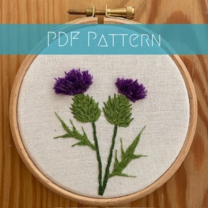 May include: Embroidered purple thistle flowers with green stems and leaves on a white fabric background in a wooden embroidery hoop.