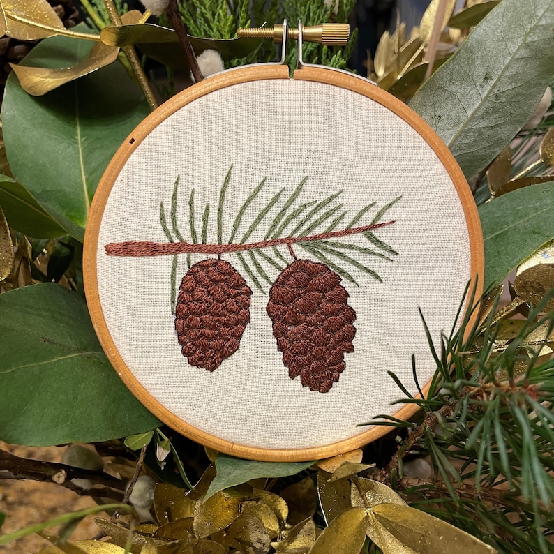 May include: A round embroidery hoop featuring a detailed needlepoint design of two brown pinecones hanging from a branch with green needles. The embroidery is set against a cream-colored fabric, framed by a wooden hoop.