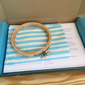 May include: A wooden embroidery hoop with a metal clasp and screw, resting on blue and white striped tissue paper inside a cardboard box. The tissue paper has the text "EMBELLISHED ELEPHANT" and "Enjoy! Sarah".