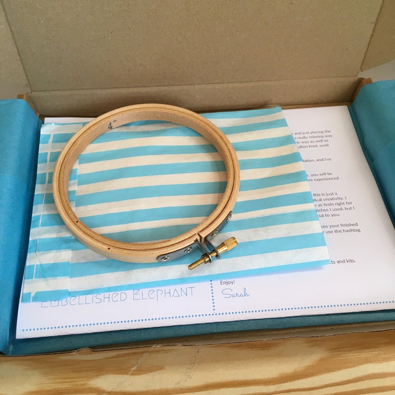 May include: A wooden embroidery hoop with a metal clasp and screw, resting on blue and white striped tissue paper inside a cardboard box. The tissue paper has the text "EMBELLISHED ELEPHANT" and "Enjoy! Sarah".