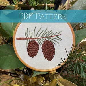May include: Embroidered pine cone and branch design in a wooden embroidery hoop. The pine cone is brown and the branch is green.