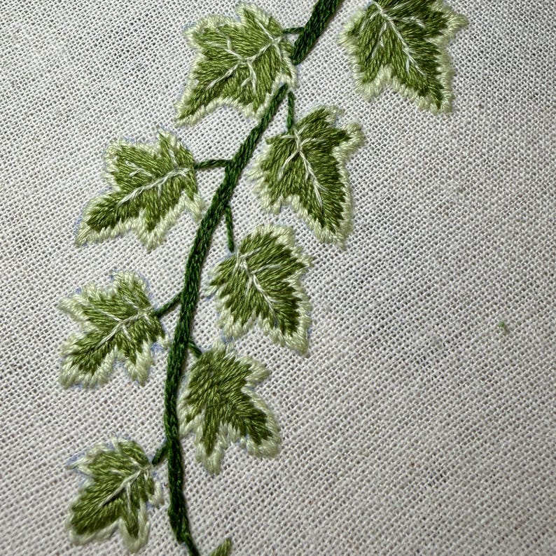 K&ouml;nnte beinhalten: Eine handgestickte Ranke mit mehreren spitzen Bl&auml;ttern in verschiedenen Gr&uuml;nt&ouml;nen, die auf einen strukturierten wei&szlig;en Stoffgrund gestickt sind. Das Design zeigt einen zentralen St&auml;ngel mit abzweigenden Bl&auml;ttern, wobei Plattstich-Sticktechniken verwendet werden.