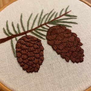 May include: Hand-stitched embroidery of two brown pine cones and green pine needles on a brown branch. The embroidery is set against a beige fabric background, framed by a wooden hoop. The design showcases a nature-inspired theme.