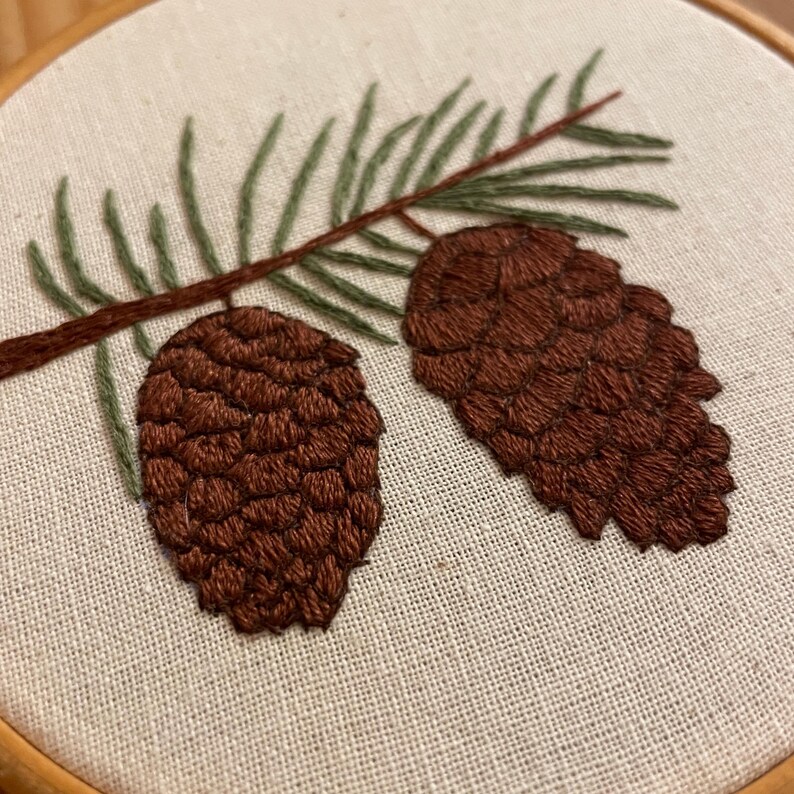 May include: Hand-stitched embroidery of two brown pine cones and green pine needles on a brown branch. The embroidery is set against a beige fabric background, framed by a wooden hoop. The design showcases a nature-inspired theme.