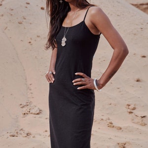 Può includere: Una donna indossa un lungo abito nero senza maniche con una collana con ciondolo d'argento e un braccialetto d'argento. È in piedi in un ambiente desertico.