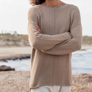 Peut inclure: Un pull en tricot beige à col bateau et manches longues. Le pull présente une couture verticale sur le devant. La personne qui le porte a de longs cheveux bruns ondulés et se tient sur une plage, les bras croisés.