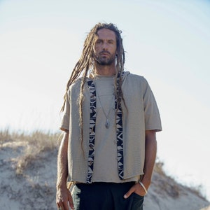 Op de afbeelding: Een man met lange dreadlocks draagt een beige linnen shirt met een zwart-witte gepatroneerde rand. Hij draagt ook een donkerblauwe korte broek en een zilveren ketting met een hanger.