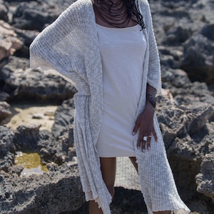 May include: A woman wearing a light gray, open-front, long knit cardigan over a light beige dress. The cardigan has a loose, flowing design. She is also wearing a necklace and rings. The background is a rocky, outdoor setting.