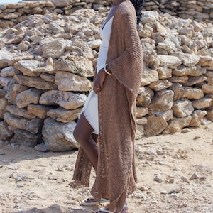 Könnte beinhalten: Eine Frau trägt einen langen, braunen Cardigan mit offenem Schnitt über einem weißen Kleid. Der Cardigan hat weite Ärmel und ein fließendes Design. Die Frau steht im Freien in der Nähe einer Steinstruktur auf sandigem Boden.