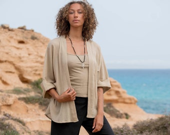 Puede incluir: Una mujer con una chaqueta de lino beige, un top sin mangas a juego y pantalones negros. También lleva un collar con un colgante de cruz y sandalias. El fondo muestra un paisaje costero con un acantilado rocoso y el mar.