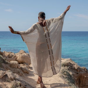 Op de afbeelding: Een vrouw draagt een lange, vloeiende, beige linnen kaftan met bruine en gouden versieringen. Ze staat op een rotsachtige klif met uitzicht op de zee.