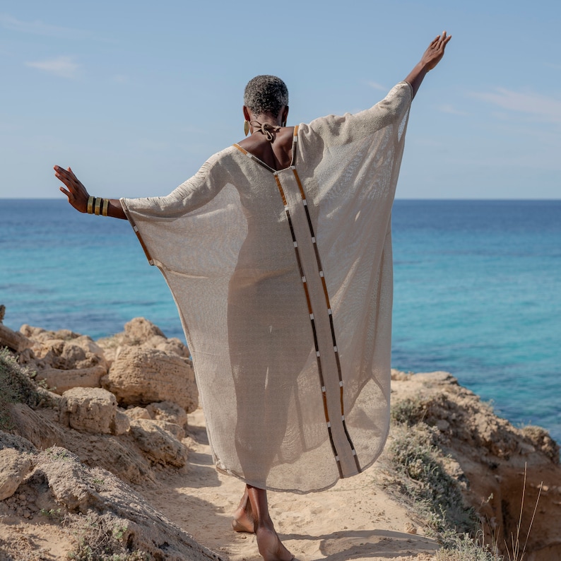 Op de afbeelding: Een vrouw draagt een lange, vloeiende, beige linnen kaftan met bruine en gouden versieringen. Ze staat op een rotsachtige klif met uitzicht op de zee.