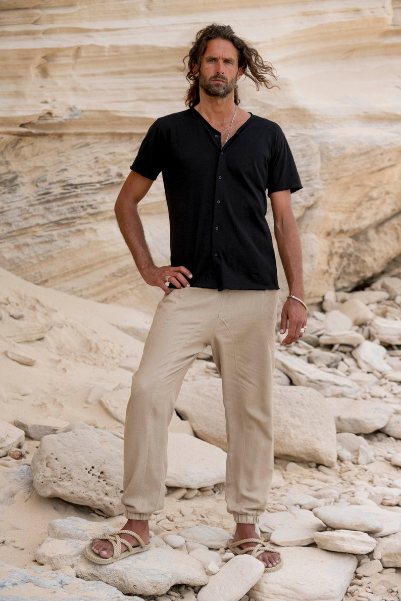 May include: A man wearing a black short-sleeved shirt with a button-up collar and beige trousers. He is standing on a rocky surface.
