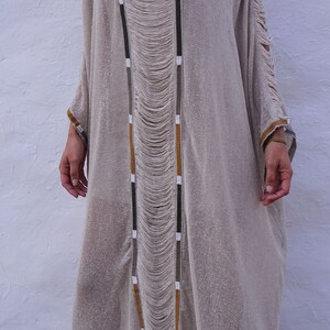 Op de afbeelding: Een lange, beige, gehaakte jurk met bruine, witte en zwarte bies. De jurk heeft een losse pasvorm en een vloeiende silhouet. De jurk is gemaakt van een lichtgewicht, ademende stof.