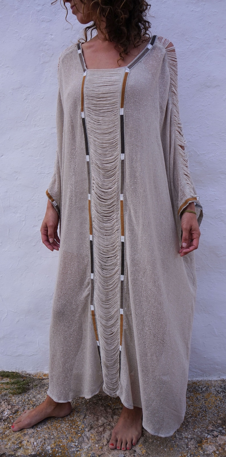 Op de afbeelding: Een lange, beige, gehaakte jurk met bruine, witte en zwarte bies. De jurk heeft een losse pasvorm en een vloeiende silhouet. De jurk is gemaakt van een lichtgewicht, ademende stof.