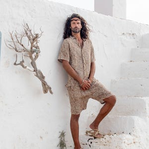 May include: A matching beige patterned shirt and shorts set. The short-sleeved button-down shirt and shorts have a repeating geometric design. The person is wearing sandals and standing on white steps.