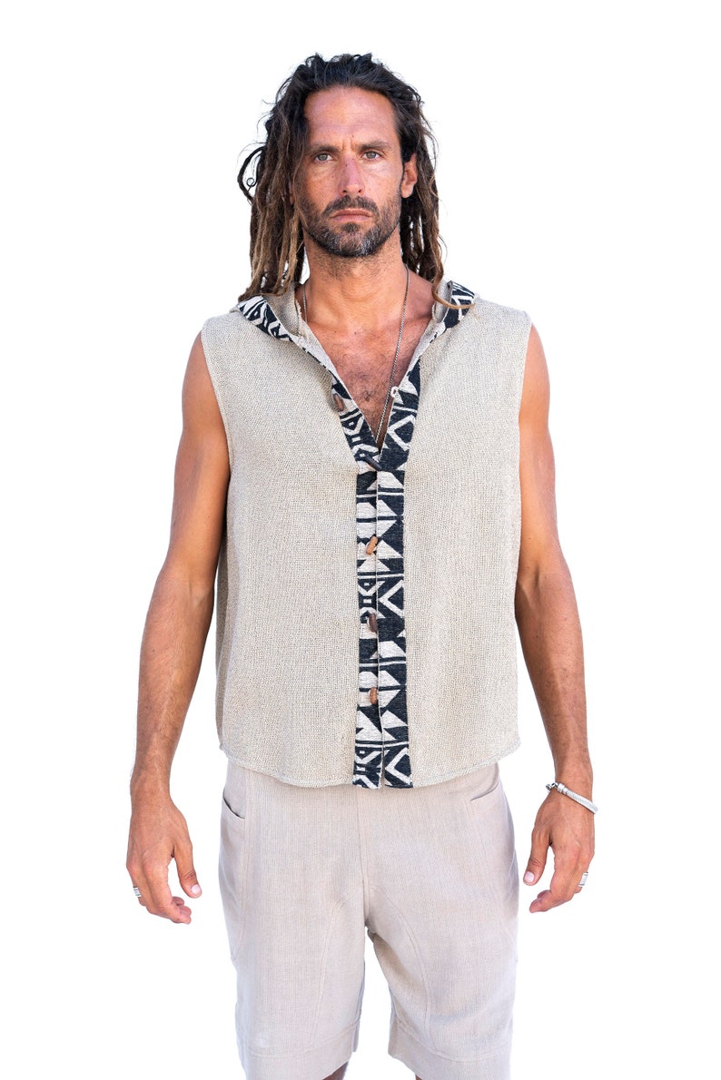 Op de afbeelding: Een mouwloos hooded shirt met een zwart-wit geometrisch patroon op de voorkant. Het shirt is gemaakt van een licht beige stof en heeft een knoopsluiting.
