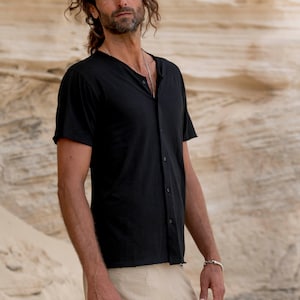 May include: A man wearing a black, button-down shirt with a v-neck and a silver necklace. He is wearing beige trousers and a silver bracelet on his left wrist.