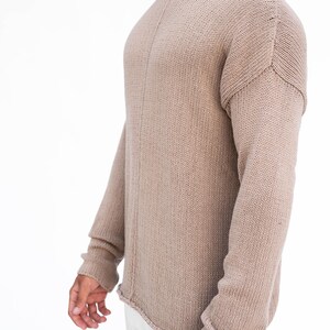 May include: A beige knitted jumper with long sleeves. The jumper features a boat neck and a slightly distressed hemline. The sleeves have ribbed cuffs. The garment is made of a soft, textured material.
