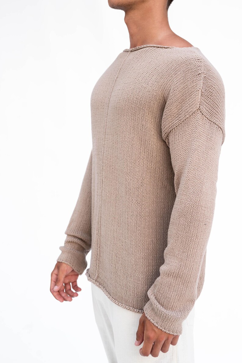May include: A beige knitted jumper with long sleeves. The jumper features a boat neck and a slightly distressed hemline. The sleeves have ribbed cuffs. The garment is made of a soft, textured material.