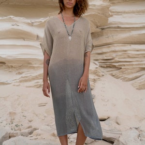 Puede incluir: Una mujer con un vestido largo de punto de color marrón claro y gris degradado con un collar de plata con una cruz. Está de pie sobre una roca en un paisaje desértico.