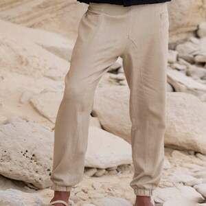 May include: A pair of beige linen trousers with a drawstring waist and elasticated cuffs. The trousers are being worn by a person standing on a rocky surface.
