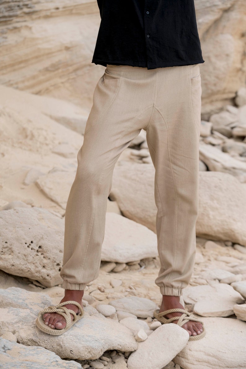 May include: A pair of beige linen trousers with a drawstring waist and elasticated cuffs. The trousers are being worn by a person standing on a rocky surface.