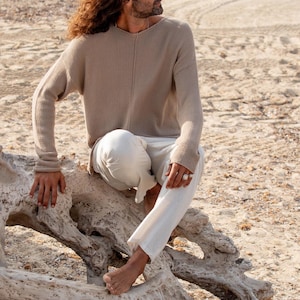 May include: A person wearing a beige knit sweater and white trousers sits on a weathered log on a sandy beach. The sweater has a v-neck and long sleeves. The trousers are loose-fitting. The person is wearing a ring.
