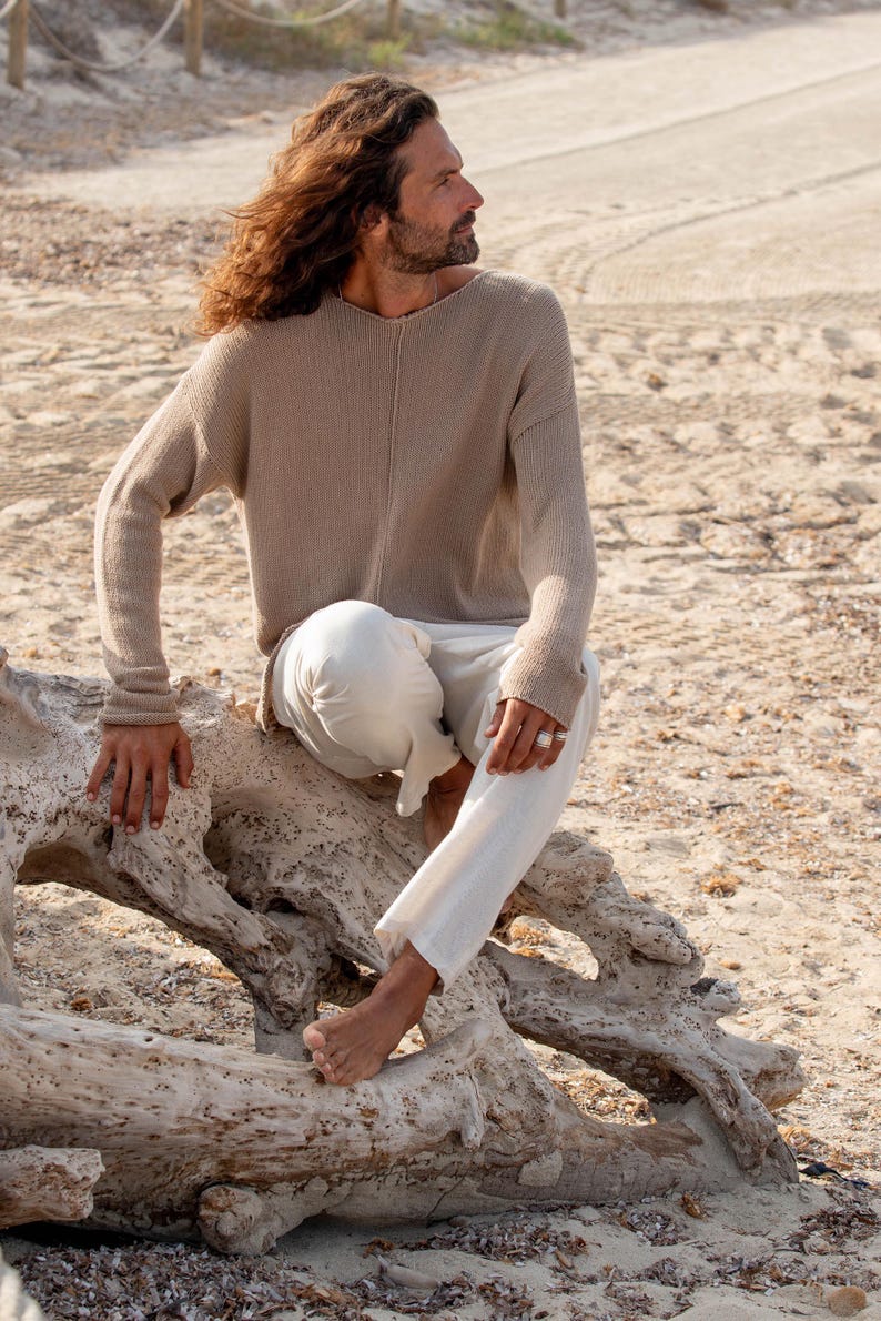 May include: A person wearing a beige knit sweater and white trousers sits on a weathered log on a sandy beach. The sweater has a v-neck and long sleeves. The trousers are loose-fitting. The person is wearing a ring.