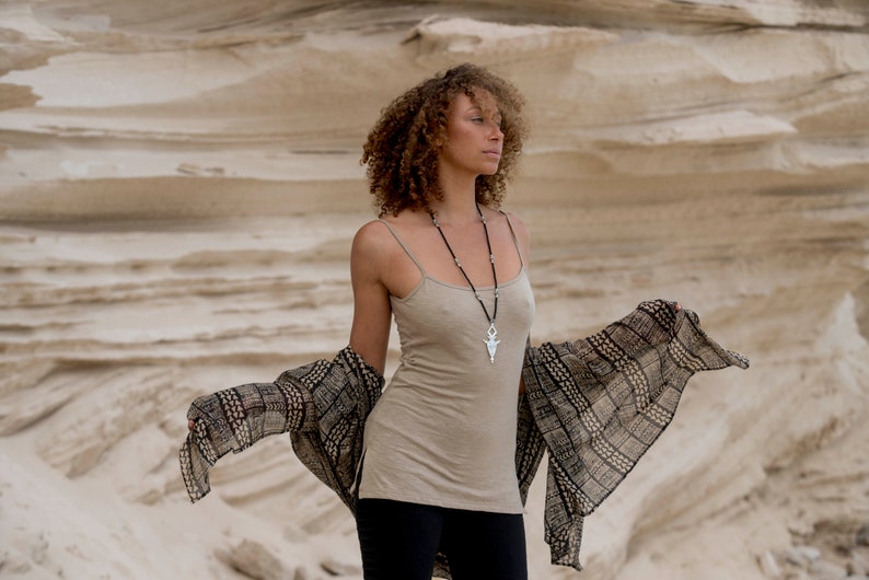 Op de afbeelding: Een vrouw met krullend bruin haar draagt een beige tanktop, een zwarte broek en een bruin-witte gestreepte sjaal. Ze draagt een zilveren ketting met een hanger.