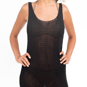 Op de afbeelding: Zwarte gebreide tankjurk met een ronde hals. De jurk is mouwloos en heeft een losse, open weefsel. Het model draagt een beha eronder. De jurk is knielang en heeft een kleine split aan de zijkant.