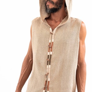May include: A sleeveless hooded vest with a beige linen fabric and a brown, white, and green trim. The vest has a button closure and a rope tie at the bottom.