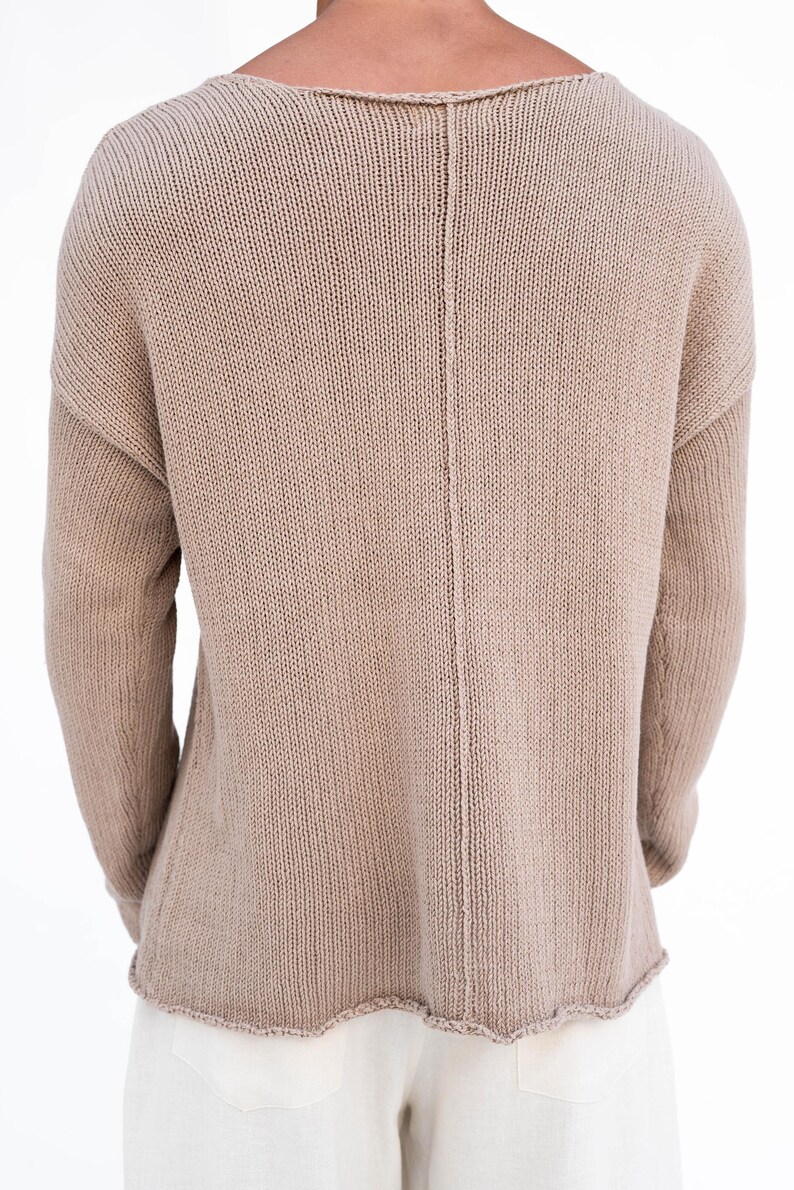 May include: A beige knitted sweater with a wide neckline and long sleeves. The back of the sweater has a vertical seam down the centre. The bottom hem has a scalloped edge. The sweater is made of a soft, textured knit fabric.