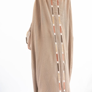 Op de afbeelding: Een lange, losse, beige gebreide jurk met een V-hals en een decoratieve bies aan de achterkant. De bies is gemaakt van bruine, oranje en witte strepen.