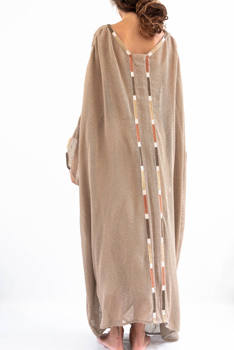 Op de afbeelding: Een lange, losse, beige gebreide jurk met een V-hals en een decoratieve bies aan de achterkant. De bies is gemaakt van bruine, oranje en witte strepen.