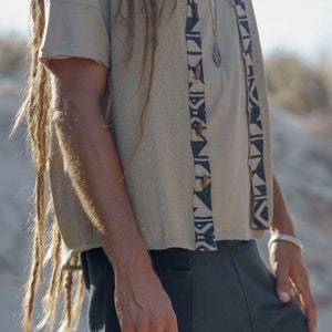 Op de afbeelding: Een persoon draagt een beige gebreide vest met zwarte en witte geometrische patronen, een beige T-shirt en een donkergroene broek. Het vest heeft houten knopen en de persoon draagt een zilveren ketting met een hanger.