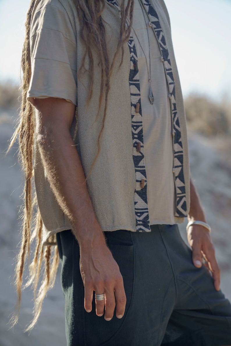 Op de afbeelding: Een persoon draagt een beige gebreide vest met zwarte en witte geometrische patronen, een beige T-shirt en een donkergroene broek. Het vest heeft houten knopen en de persoon draagt een zilveren ketting met een hanger.