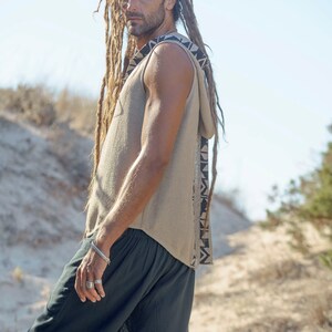 Op de afbeelding: Een man met lange dreadlocks draagt een mouwloos hooded top met een geometrisch patroon aan de zijkant. Hij draagt ook een zwarte short.