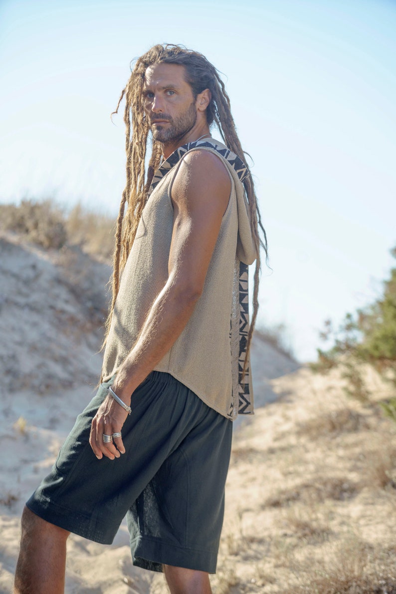 Op de afbeelding: Een man met lange dreadlocks draagt een mouwloos hooded top met een geometrisch patroon aan de zijkant. Hij draagt ook een zwarte short.