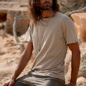May include: A man wearing a light brown t-shirt with a silver chain necklace and a pendant. He is also wearing olive green shorts and a silver bracelet on his wrist.