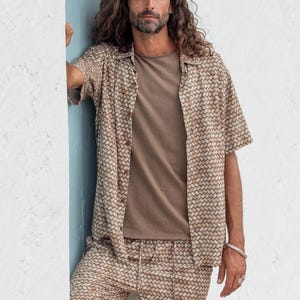 May include: A matching beige shirt and shorts set with a woven pattern. The short-sleeved shirt is buttoned and worn over a solid brown t-shirt. The shorts have a drawstring waist. The outfit is casual and suitable for warm weather.
