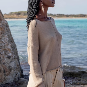 Puede incluir: Una persona con un jersey de punto beige y pantalones a juego junto al mar. El jersey tiene un corte holgado y mangas largas. Los pantalones son de cintura alta y de corte holgado. Lleva pendientes y una pulsera plateados.