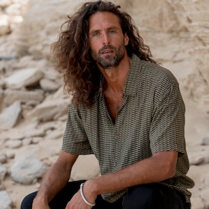 Può includere: Un uomo con lunghi capelli castani indossa una camicia a maniche corte con motivo nero e oro e pantaloni neri. È seduto su una roccia con uno sfondo bianco.