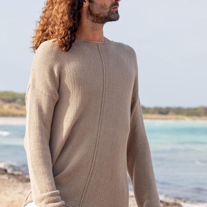 May include: A beige knitted jumper with long sleeves. The jumper has a relaxed fit and a seam detail down the centre. The person is wearing white trousers. The background is a beach with blue water and a clear sky.
