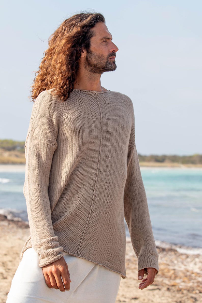 May include: A beige knitted jumper with long sleeves. The jumper has a relaxed fit and a seam detail down the centre. The person is wearing white trousers. The background is a beach with blue water and a clear sky.