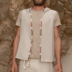 May include: A beige hooded vest with brown and white trim. The vest has a button closure and is made of a soft, textured fabric.