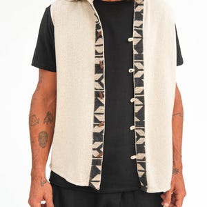 Op de afbeelding: Een mouwloos vest met capuchon met een geometrisch patroon aan de voorkant. Het vest is gemaakt van licht beige stof en heeft een zwart-wit geometrisch patroon aan de voorkant. Het vest is aan de voorkant met knopen gesloten.