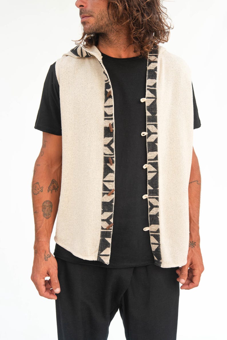 Op de afbeelding: Een mouwloos vest met capuchon met een geometrisch patroon aan de voorkant. Het vest is gemaakt van licht beige stof en heeft een zwart-wit geometrisch patroon aan de voorkant. Het vest is aan de voorkant met knopen gesloten.